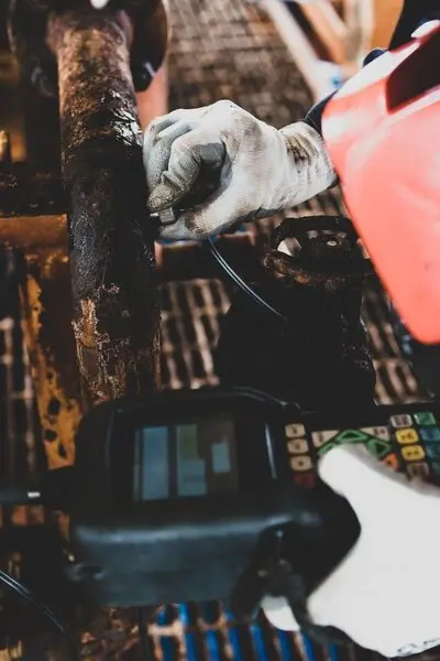 A worker inspects heavily corroded industrial pipes. An ultrasonic tool tests the remaining metal thickness.