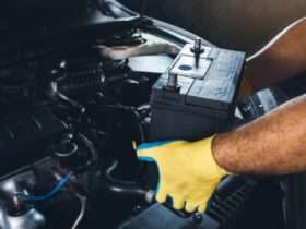 A person's yellow-gloved hands holding a black car battery above an open vehicle engine compartment with dark parts.