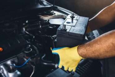 A person's yellow-gloved hands holding a black car battery above an open vehicle engine compartment with dark parts.