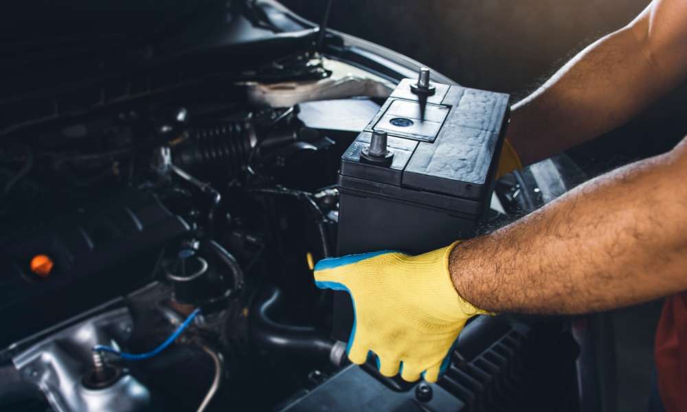 A person's yellow-gloved hands holding a black car battery above an open vehicle engine compartment with dark parts.