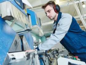 An operator wearing ear protection and safety glasses stands next to the side of a plastic fabrication machine.