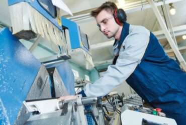 An operator wearing ear protection and safety glasses stands next to the side of a plastic fabrication machine.