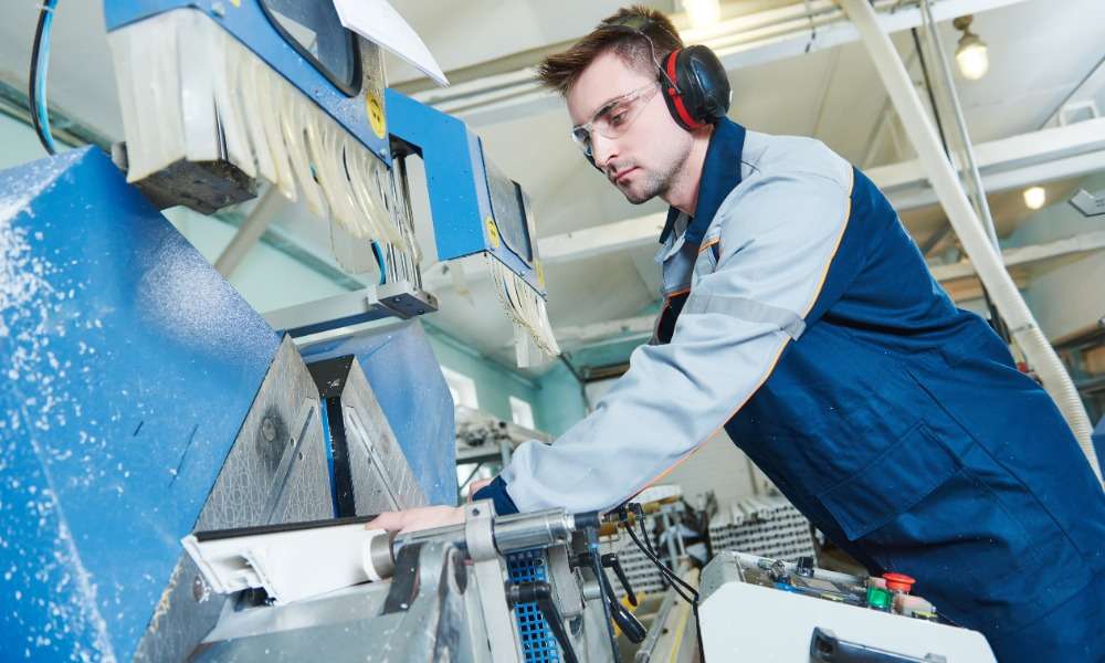 An operator wearing ear protection and safety glasses stands next to the side of a plastic fabrication machine.