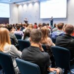 A large crowd of attendees sitting in dark blue chairs in front of a speaker inside a massive, bright room.