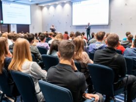 A large crowd of attendees sitting in dark blue chairs in front of a speaker inside a massive, bright room.