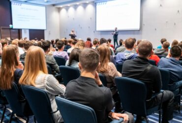 A large crowd of attendees sitting in dark blue chairs in front of a speaker inside a massive, bright room.