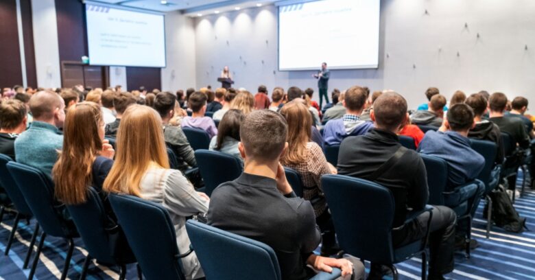 A large crowd of attendees sitting in dark blue chairs in front of a speaker inside a massive, bright room.