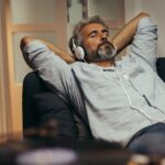 A man lies on a couch with his hands behind his head and his eyes closed. He wears over-the-ear headphones.