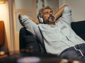 A man lies on a couch with his hands behind his head and his eyes closed. He wears over-the-ear headphones.