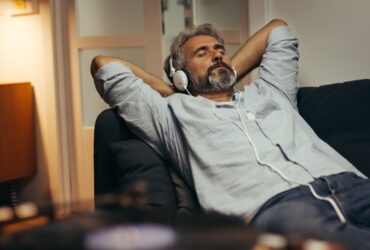 A man lies on a couch with his hands behind his head and his eyes closed. He wears over-the-ear headphones.