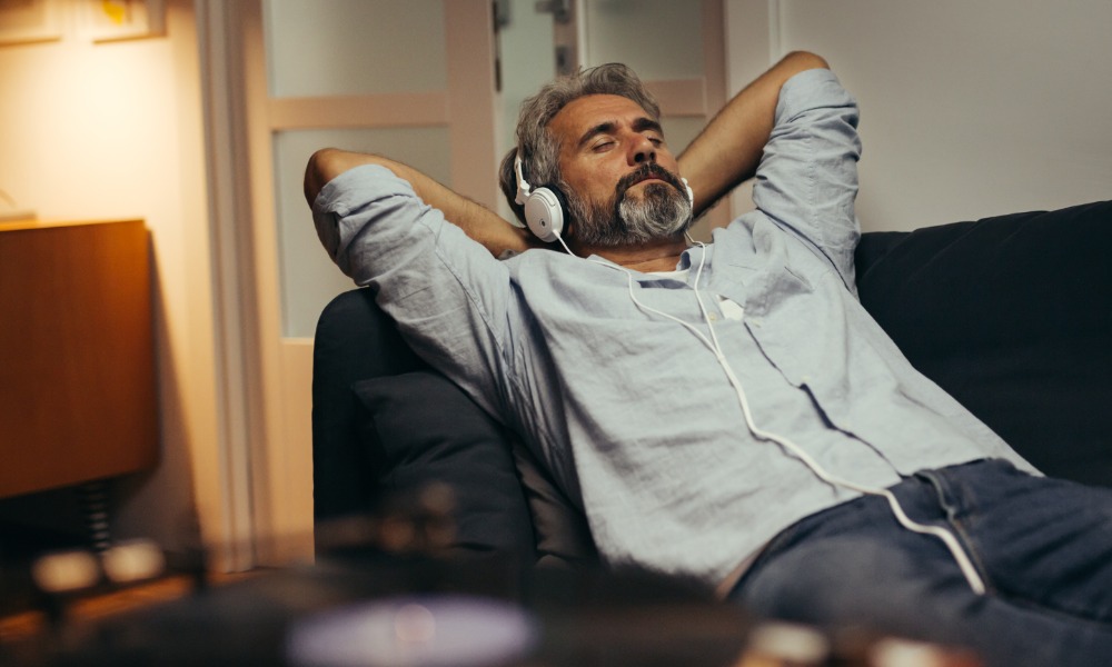 A man lies on a couch with his hands behind his head and his eyes closed. He wears over-the-ear headphones.