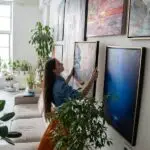 A person hanging art on a wall full of other artwork. Sunlight shines into the home through a nearby window.