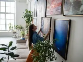 A person hanging art on a wall full of other artwork. Sunlight shines into the home through a nearby window.