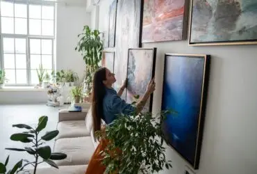 A person hanging art on a wall full of other artwork. Sunlight shines into the home through a nearby window.
