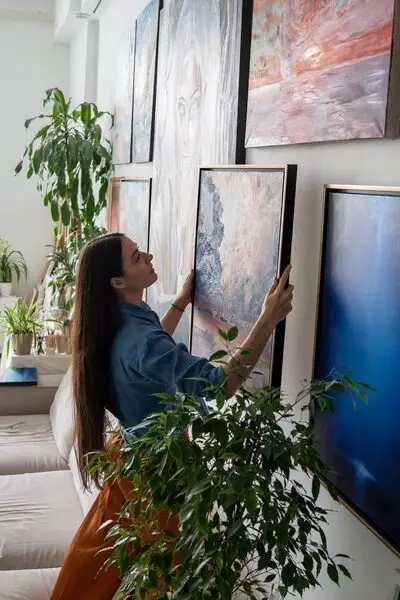 A person hanging art on a wall full of other artwork. Sunlight shines into the home through a nearby window.