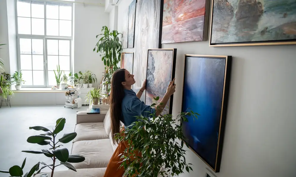 A person hanging art on a wall full of other artwork. Sunlight shines into the home through a nearby window.
