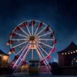 A massive brightly lit ferris wheel in the middle of a carnival at nighttime. The carnival is empty.