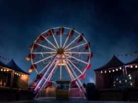 A massive brightly lit ferris wheel in the middle of a carnival at nighttime. The carnival is empty.
