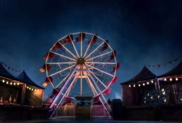 A massive brightly lit ferris wheel in the middle of a carnival at nighttime. The carnival is empty.