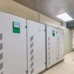 Two large industrial voltage stabilizers with digital controls installed against a white wall in a factory.