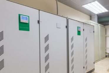 Two large industrial voltage stabilizers with digital controls installed against a white wall in a factory.