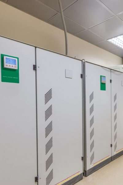 Two large industrial voltage stabilizers with digital controls installed against a white wall in a factory.