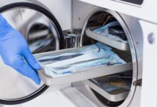 Gloved hand loads a tray of packaged medical instruments into an open stainless steel autoclave for sterilization.