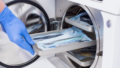 Gloved hand loads a tray of packaged medical instruments into an open stainless steel autoclave for sterilization.