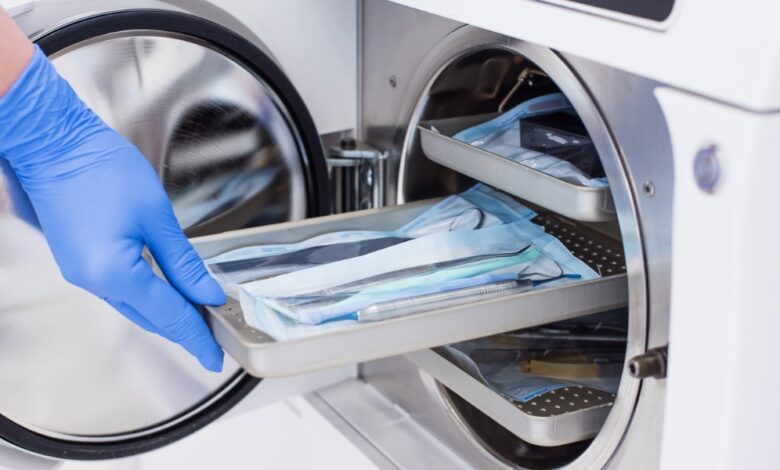 Gloved hand loads a tray of packaged medical instruments into an open stainless steel autoclave for sterilization.