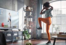 A woman in athletic clothes exercising on a mat in a living room while looking toward a television screen.