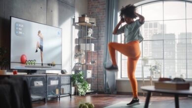 A woman in athletic clothes exercising on a mat in a living room while looking toward a television screen.