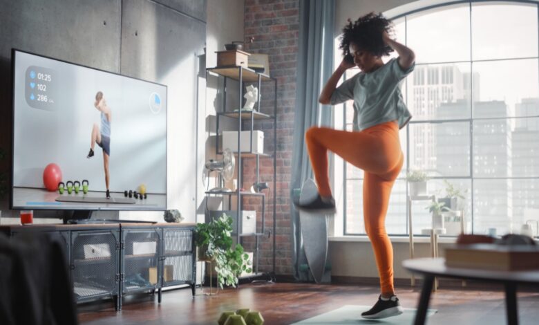 A woman in athletic clothes exercising on a mat in a living room while looking toward a television screen.