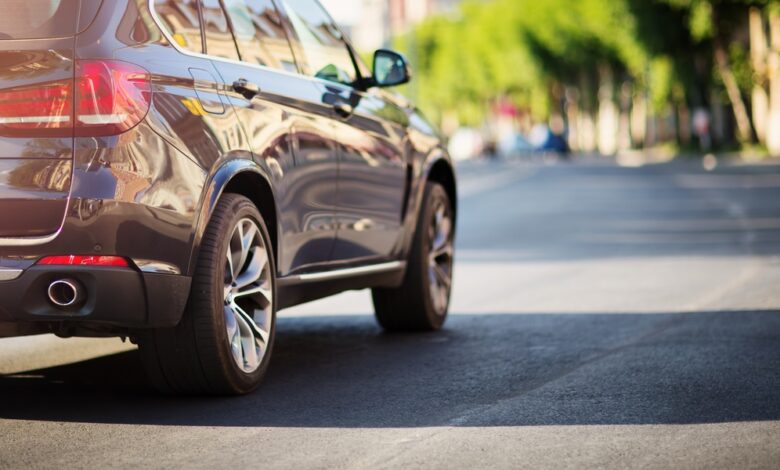 A dark SUV on a sunny city street, with trees lining the road and blurred vehicles in the distance ahead.