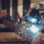 A person wearing a welding helmet working on a metal beam as bright sparks and smoke fill an industrial shop.