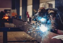 A person wearing a welding helmet working on a metal beam as bright sparks and smoke fill an industrial shop.