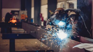 A person wearing a welding helmet working on a metal beam as bright sparks and smoke fill an industrial shop.