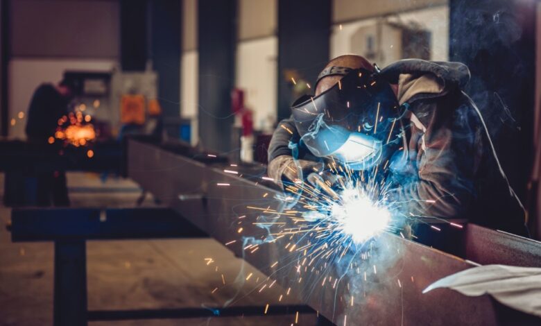 A person wearing a welding helmet working on a metal beam as bright sparks and smoke fill an industrial shop.