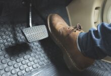 A person wearing a brown leather boot and blue jeans presses a gray brake pedal inside a car with black rubber floor mats.