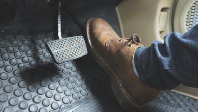 A person wearing a brown leather boot and blue jeans presses a gray brake pedal inside a car with black rubber floor mats.