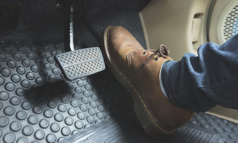 A person wearing a brown leather boot and blue jeans presses a gray brake pedal inside a car with black rubber floor mats.