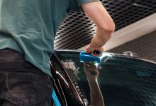 A person in a green shirt using a blue squeegee on a wet car windshield inside an automotive workshop.