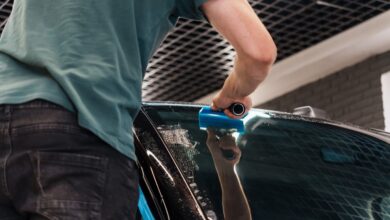 A person in a green shirt using a blue squeegee on a wet car windshield inside an automotive workshop.