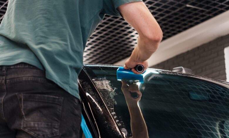 A person in a green shirt using a blue squeegee on a wet car windshield inside an automotive workshop.