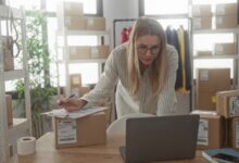 A woman checking shipping paperwork beside a laptop and labeled packages in a small business workspace.