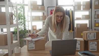 A woman checking shipping paperwork beside a laptop and labeled packages in a small business workspace.