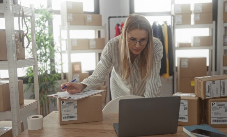 A woman checking shipping paperwork beside a laptop and labeled packages in a small business workspace.