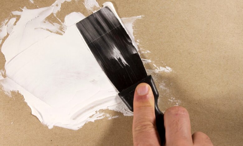 A close-up of a beige wall. A person uses a putty knife to spread spackling paste over an imperfection.