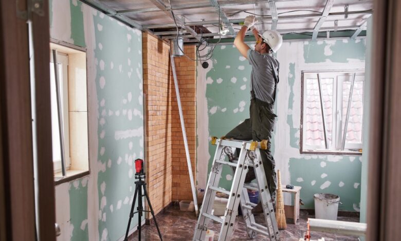 A person stands on a stepladder attaching plasterboard to a ceiling inside an unfinished room, with equipment nearby.
