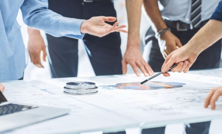 A group of coworkers gathered around a table, reviewing plans while one person points at a colorful diagram.
