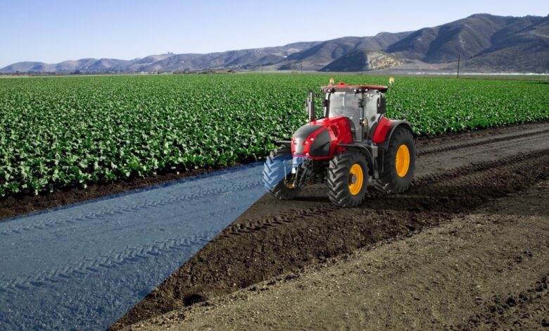 A red tractor driving along a dirt path beside green crop rows, with a blue scanning beam across the soil near mountains.